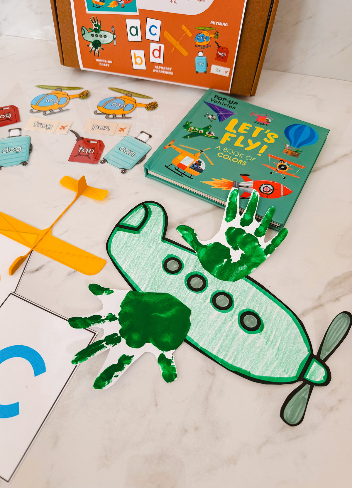 Children's educational materials including a book titled 'Let's Fly' and handprint airplane craft on a light surface.