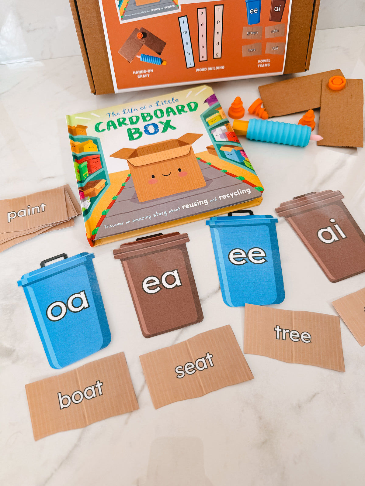 Educational materials including a cardboard box, colorful bins, and letter cards on a white surface.