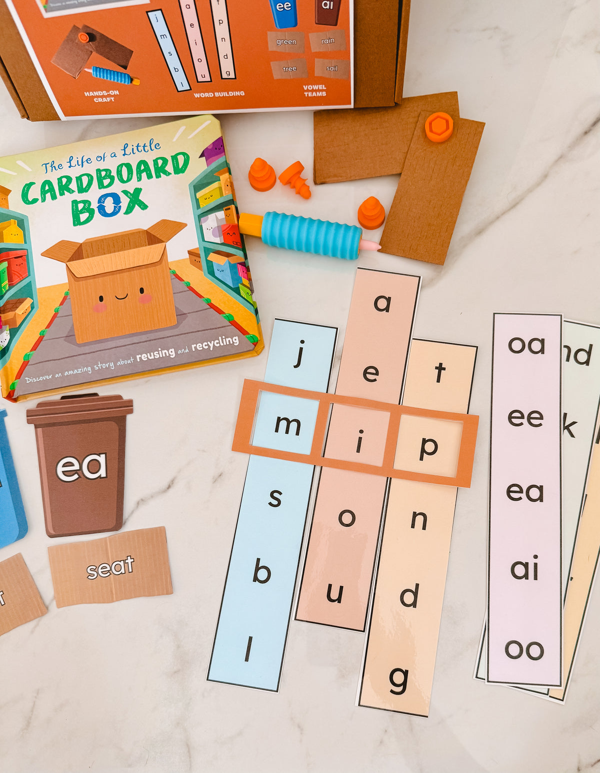 Educational word cards with letter tiles on a marble surface, featuring a cardboard box product.