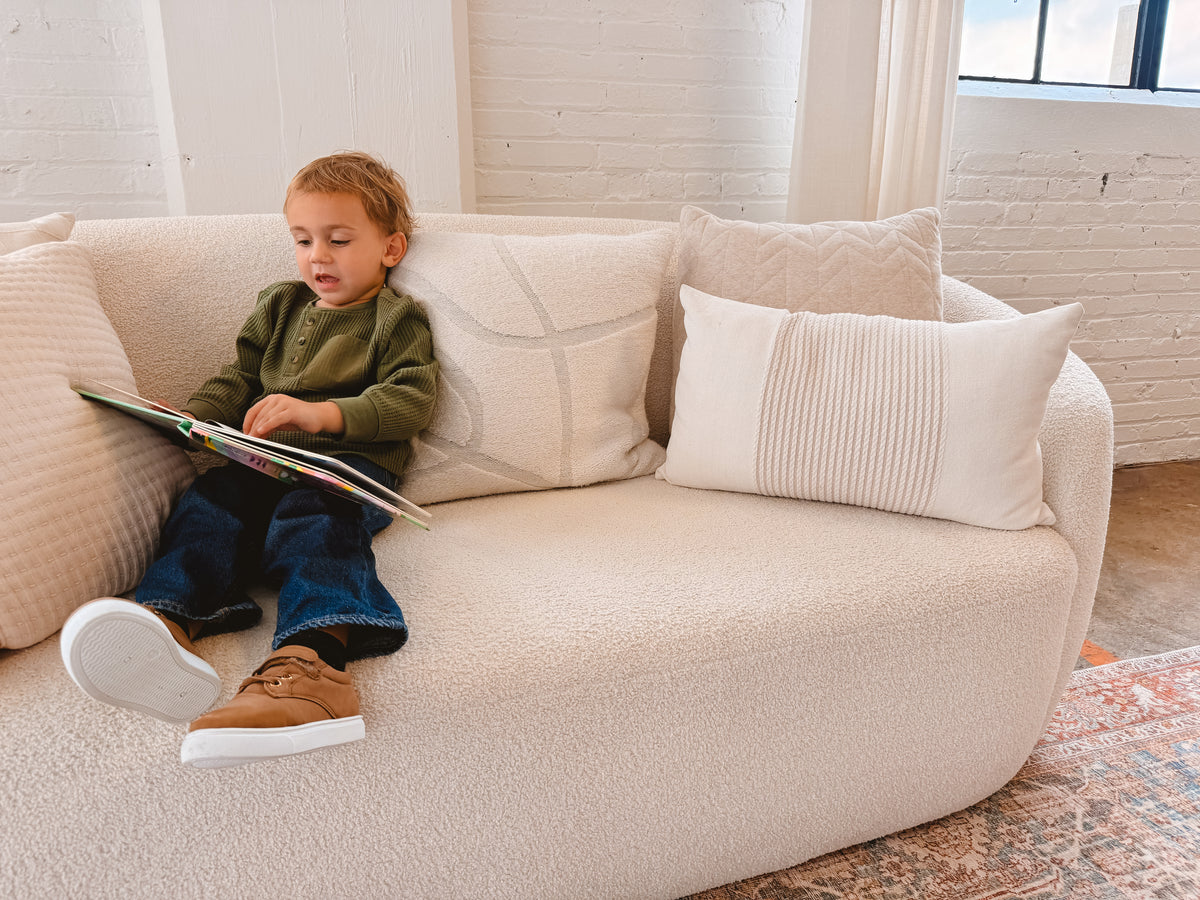 A young child sits on a cream boucle sofa reading a book, with decorative throw pillows in a bright, modern room with white brick walls.